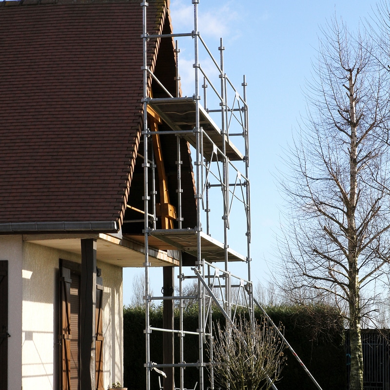 Échafaudage et Travaux de Rénovation sur Maison Traditionnelle Échafaudage métallique monté contre le pignon d'une maison à colombages, près d'un toit en tuiles rouges, sous un ciel bleu clair.