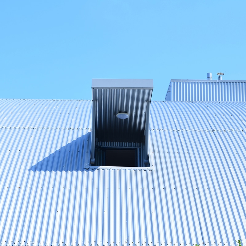 Toiture en tôle ondulée arquée avec lucarne et lumière Toiture arquée en tôle ondulée grise, avec une lucarne ouverte projetant une ombre nette sous un ciel bleu clair.